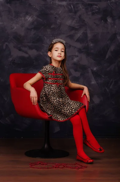 A szép kislány gyönyörű ruha piros karosszékben ül rajta. Ő visel piros maskara farsangi maszk. Stúdió felvétel. — Stock Fotó