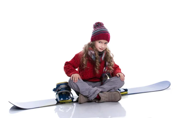 Petite fille souriante à la coiffure bouclée portant un pull tricoté, une écharpe, un chapeau et des gants assis sur un snowboard bleu, isolé sur blanc. Vêtements d'hiver et concept sportif . — Photo