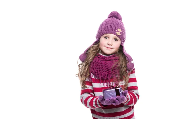 Nettes lächelndes kleines Mädchen mit lila gestricktem Schal, Mütze und Handschuhen, das Weihnachtsgeschenk isoliert auf weißem Hintergrund hält. Winterkleidung und Weihnachtskonzept. — Stockfoto