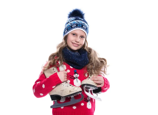 Ganska leende liten flicka med krullig frisyr bär stickad tröja, halsduk och hatt med skridskor isolerad på vit bakgrund. Vinterkläder och sport koncept. — Stockfoto
