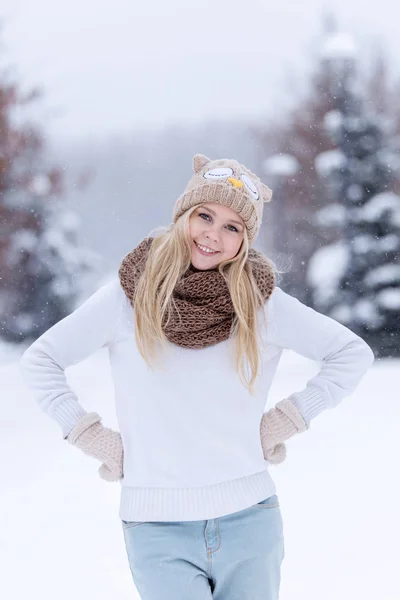 Atractiva joven rubia sonriente caminando en el bosque de invierno. Mujer bonita en invierno al aire libre. Llevando ropa de invierno. Jersey, bufanda, gorro y manoplas de punto . — Foto de Stock