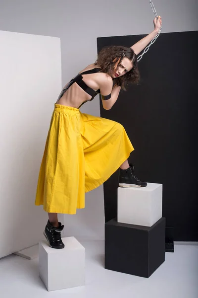 Mode foto av vacker flicka poserar i studio. Bära gula shorts, stövlar svarta. Remmar över bröstet. Håller kedjan. Lockig frisyr. — Stockfoto