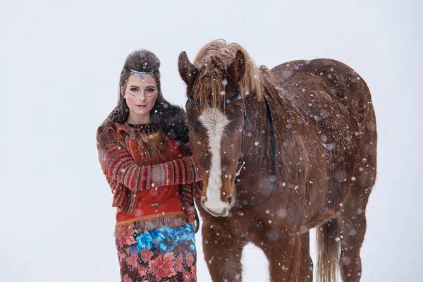 Mulher indígena nativa com maquiagem tradicional e penteado no inverno nevado. Menina bonita em vestido étnico abraçando um cavalo . — Fotografia de Stock