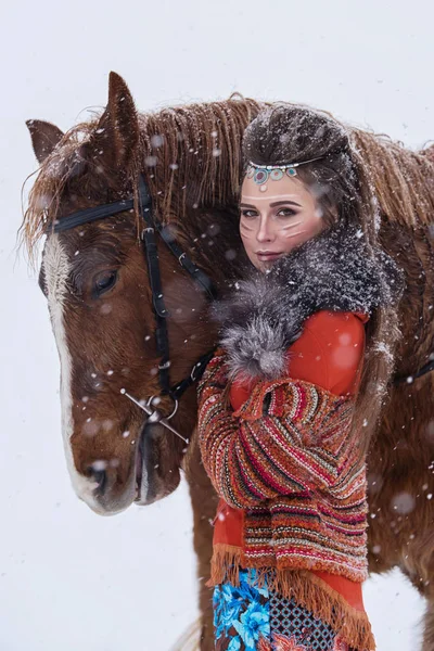 Mulher indígena nativa com maquiagem tradicional e penteado no inverno nevado. Menina bonita em vestido étnico abraçando um cavalo . — Fotografia de Stock