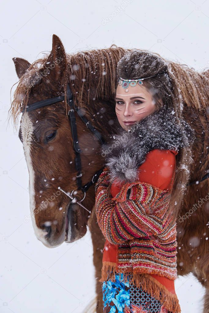 Native indian woman with traditional makeup and hairstyle in snowy winter. Beautiful girl in ethnic dress hugging a horse.
