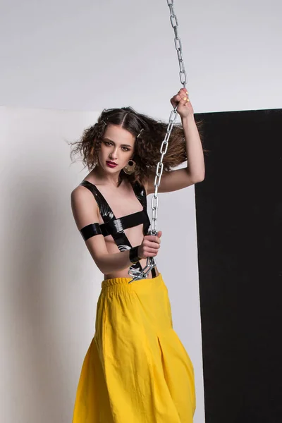 Mode foto van mooi meisje poseren in de studio. Het dragen van gele broek, laarzen zwarte. Banden over de borst. Keten te houden. Krullend kapsel. — Stockfoto