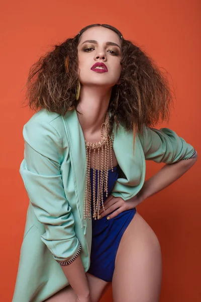 Divat fotó stúdióban szép lány. Fiatal barna nő kék fehérnemű és türkiz kabát, pózol, piros háttér. Göndör frizura. — Stock Fotó
