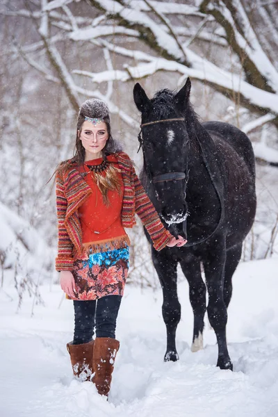 Mulher indígena nativa com maquiagem tradicional e penteado no inverno nevado. Menina bonita em vestido étnico na floresta com um cavalo . — Fotografia de Stock