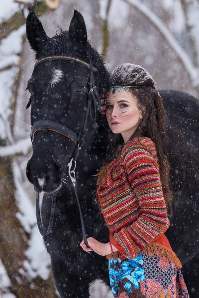 Mulher indígena nativa com maquiagem tradicional e penteado no inverno nevado. Menina bonita em vestido étnico na floresta abraçando um cavalo . — Fotografia de Stock