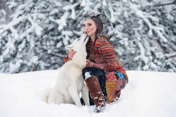 Mujer indígena nativa con maquillaje tradicional y peinado en invierno nevado. Hermosa chica en vestido étnico abrazando a un perro esquimal . — Foto de Stock