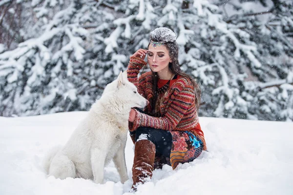 Mujer indígena nativa con maquillaje tradicional y peinado en invierno nevado. Hermosa chica en vestido étnico abrazando a un perro esquimal . — Foto de Stock