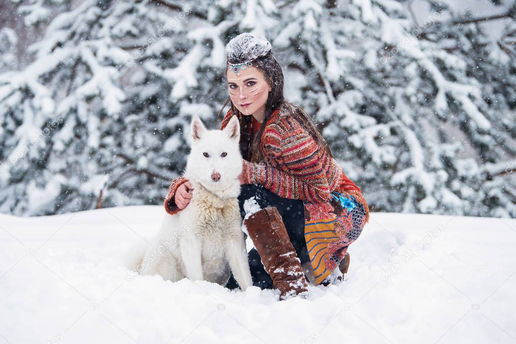 Native indian woman with traditional makeup and hairstyle in snowy winter. Beautiful girl in ethnic dress hugging an eskimo dog.
