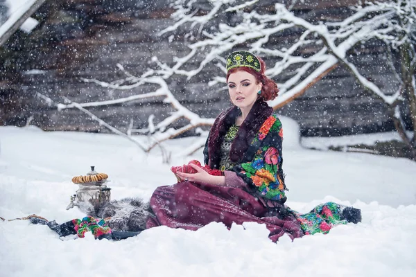 Portret van een prachtige jonge vrouw in Russische stijl jurk op een sterke vorst in een winterse besneeuwde dag met appels en Samowar. Russische model meisje in traditionele hoofdtooi. — Stockfoto