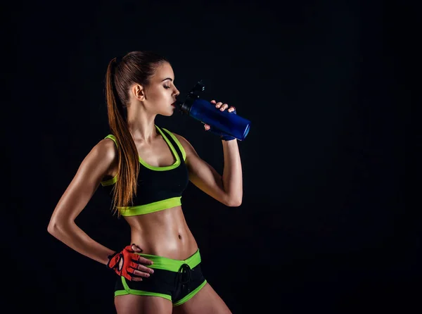 Young athletic woman in sportswear with a shaker in studio against black background. Ideal female sports figure. Fitness girl with perfect sculpted muscular and tight body. — Stock Photo, Image