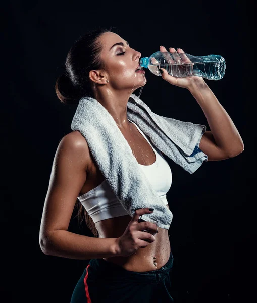 Attraente giovane donna atletica con corpo perfetto acqua potabile da una bottiglia con asciugamano intorno al collo su sfondo nero. Bella ragazza fitness rilassante dopo l'allenamento . — Foto Stock