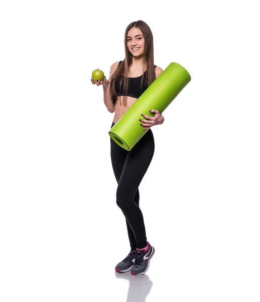 Portrait of smiling young woman holding rolled up exercise yoga mat and green apple isolated on white background. — Stock Photo, Image