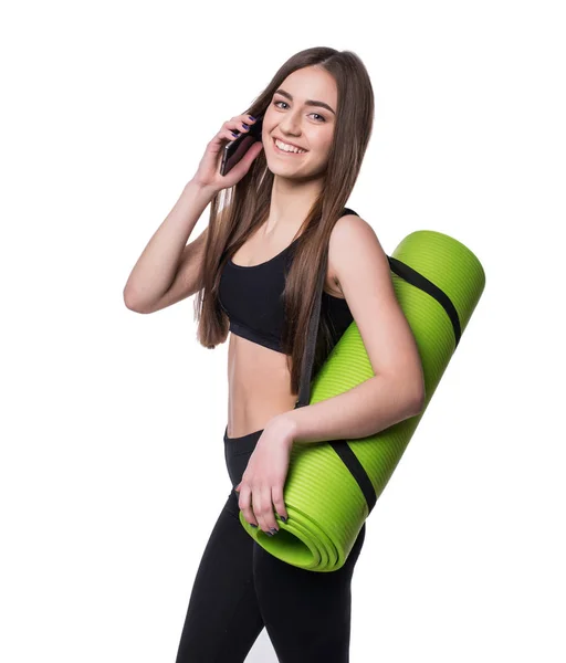 Carino giovane donna in abbigliamento sportivo con tappetino verde pronto per l'allenamento. Sorridere e parlare al telefono. Isolato su sfondo bianco . — Foto Stock