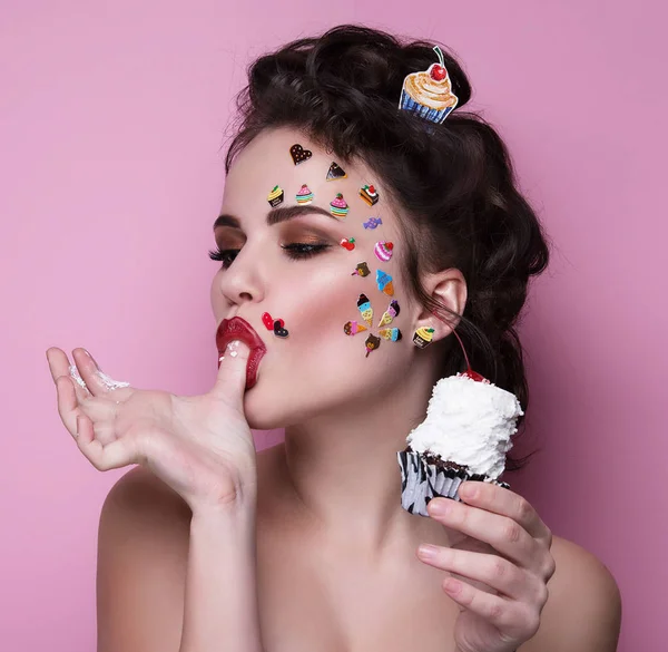 Moda Piękny Dziewczyna z luksusowych profesjonalny makijaż i emoji śmieszne naklejki przyklejone na twarzy. Młoda kobieta z cupcake w ręce. — Zdjęcie stockowe