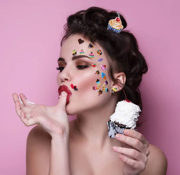 Güzel moda kız lüks profesyonel makyaj ve komik emoji çıkartmaları yüzünde yapıştırılmış. Cupcake elinde olan kadın.