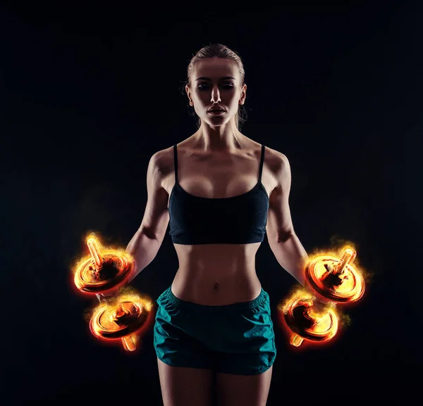 Retrato de una joven mujer de fitness en ropa deportiva haciendo ejercicio con pesas de fuego sobre fondo negro. Chica atlética sexy bronceada. Un gran cuerpo deportivo femenino . —  Fotos de Stock