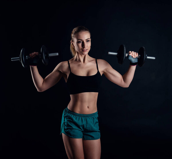 Cute athletic model girl in sportswear with dumbbells in studio against black background. Ideal female sports figure. Fitness woman with perfect sculpted muscular and tight body.