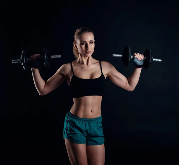 Carino ragazza modello atletico in abbigliamento sportivo con manubri in studio su sfondo nero. Figura sportiva femminile ideale. Fitness donna con perfetto scolpito corpo muscoloso e stretto . — Foto Stock