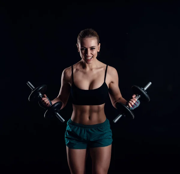 Retrato de una joven mujer de fitness en ropa deportiva haciendo ejercicio con pesas sobre fondo negro. Chica atlética sexy bronceada. Un gran cuerpo deportivo femenino . — Foto de Stock