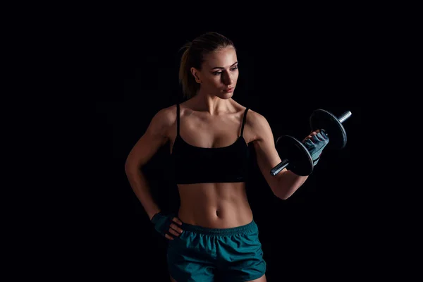Retrato de uma jovem mulher fitness em sportswear fazendo exercícios com halteres em fundo preto. Menina atlética sexy bronzeada. Um grande corpo feminino desportivo . — Fotografia de Stock