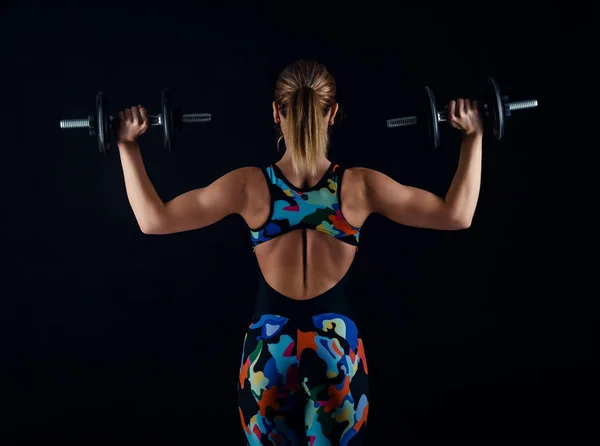 Mujer joven culturista con cuerpo musculoso fuerte perfecto que usa chándal de ropa deportiva que hace ejercicio con pesas. Vista trasera . —  Fotos de Stock