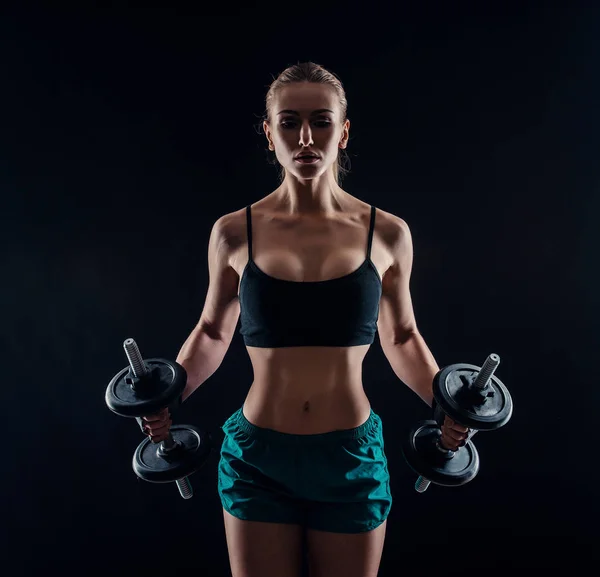 Portret van een jonge fitness vrouw in sportkleding training met halters op zwarte achtergrond. Gebruinde sexy atletische meisje. Een grote sportieve vrouwelijk lichaam. — Stockfoto