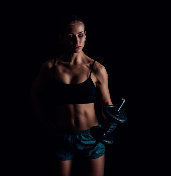 Retrato de uma jovem mulher fitness em sportswear fazendo exercícios com halteres em fundo preto. Menina atlética sexy bronzeada. Um grande corpo feminino desportivo . — Fotografia de Stock