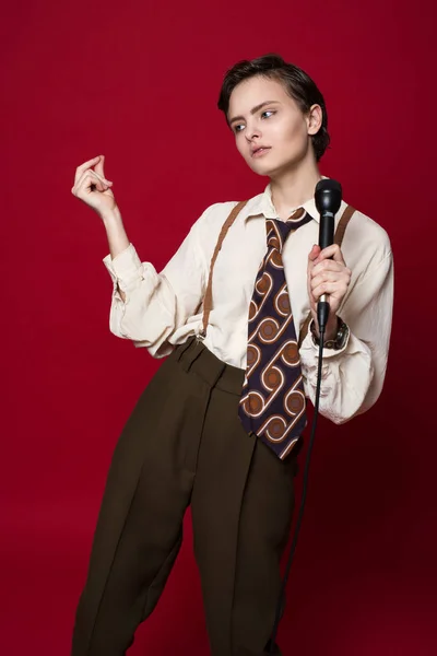 Fashionable beautiful singer girl in retro coat, tie and pants with microphone in hands posing on red background. — Stock Photo, Image