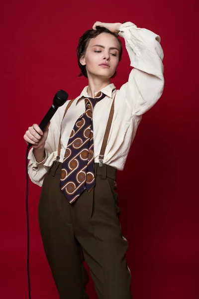 Модная красавица-певица в ретро пальто, галстуке и брюках с микрофоном в руках позирует на красном фоне . — стоковое фото