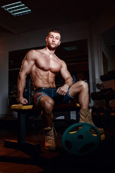 El culturista se sienta en un banco de pesas, se toma un descanso. Hombre musculoso en un lugar de entrenamiento en un gimnasio y sonriendo a la cámara . — Foto de Stock