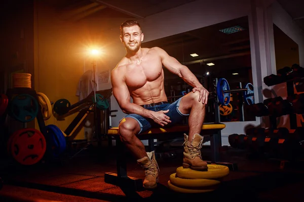 El culturista se sienta en un banco de pesas, se toma un descanso. Hombre musculoso en un lugar de entrenamiento en un gimnasio y sonriendo a la cámara . — Foto de Stock