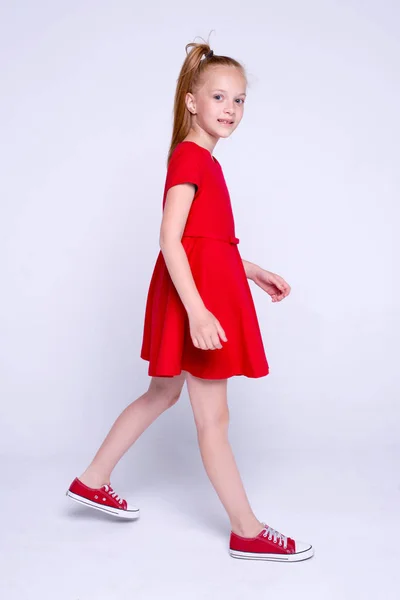 Gyönyörű vörös hajú kislány piros ruha és cipők pózol, mint a modell a fehér háttér. — Stock Fotó
