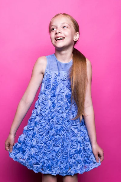 Szép kis vöröshajú lány pózol, mint a modell a rózsaszín háttér kék ruha. — Stock Fotó
