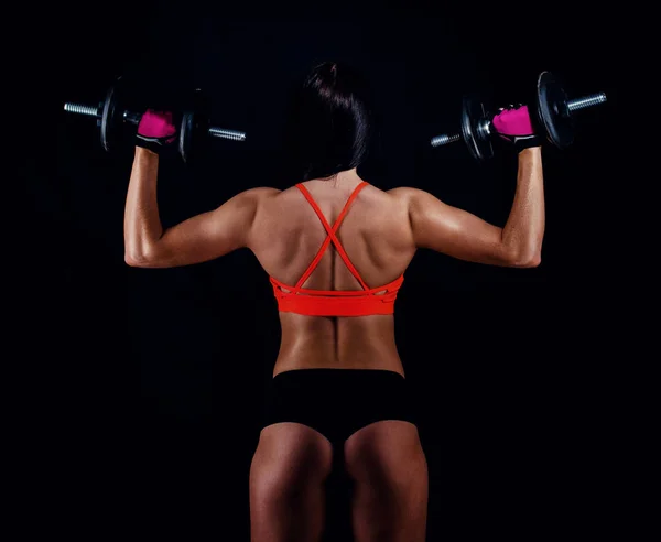 Ritratto di una giovane donna attraente in abbigliamento sportivo che fa allenamento con manubri su sfondo nero. Ragazza atletica sexy abbronzata. Un grande corpo femminile sportivo. Vista posteriore . — Foto Stock