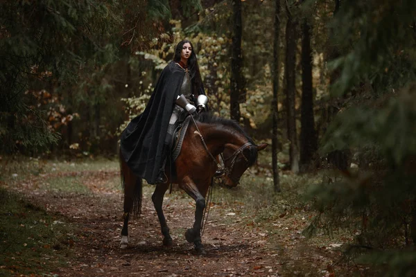 Una hermosa guerrera con una espada con cota de malla y armadura montando un caballo en un bosque misterioso . — Foto de Stock