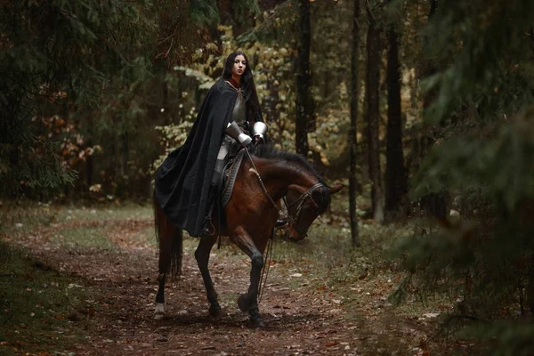 A beautiful warrior girl with a sword wearing chainmail and armor riding a horse in a mysterious forest. — Stock Photo, Image