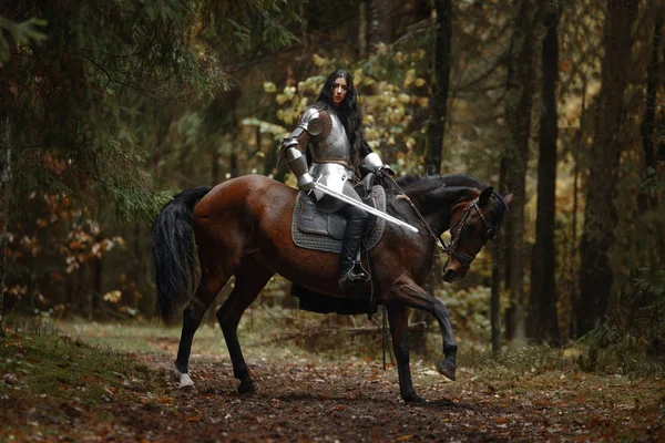 Uma bela guerreira com uma espada usando chainmail e armadura montando um cavalo em uma floresta misteriosa . — Fotografia de Stock