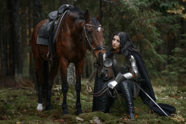 Een mooie warrior meisje met een zwaard dragen chainmail en armor met een paard in een mysterieuze bos. — Stockfoto