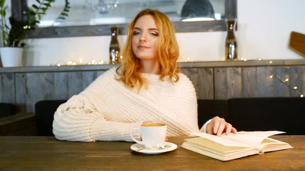 Młoda kobieta piękną jest czytanie książki w kawiarni. Uśmiecha się i cieszyć się aromatem kawy. — Wideo stockowe