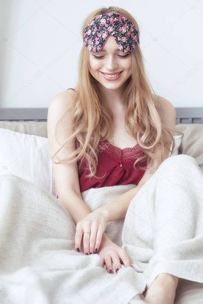 Happy smiling young woman sitting in her bed at home, wearing nighty and sleeping eye mask.