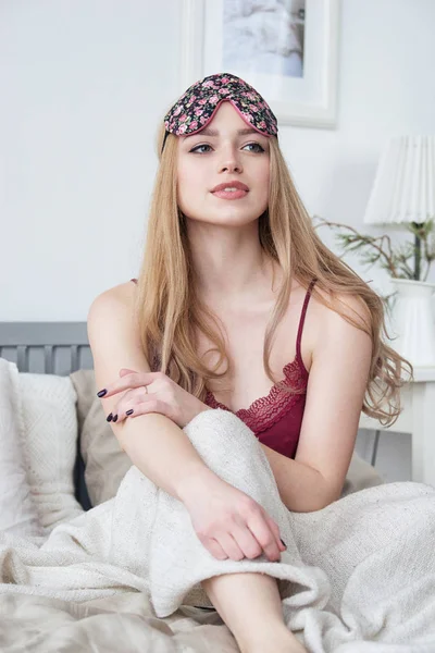 Happy smiling young woman sitting in her bed at home, wearing nighty and sleeping eye mask. — Stock Photo, Image