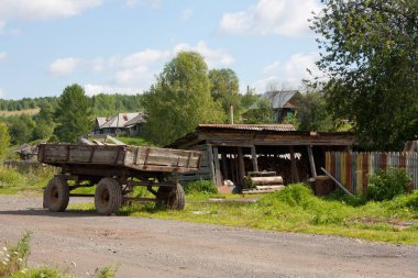 Arka planda geleneksel evler olan bir Rus kırsal manzarası. Öndeki kütük, araba, çalı ve ağaç yığını..