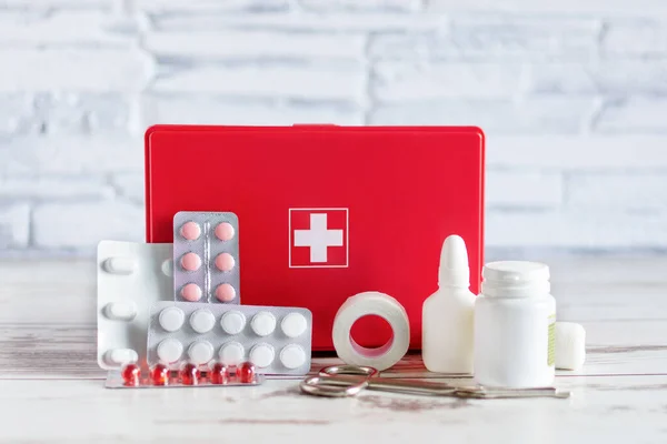 First aid kit red box with medical equipment and medications for emergency on white wooden background.