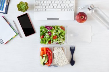 Sebzeler ve beslenme çantasında taze yeşil salata. Çalışma masasında, dizüstü bilgisayar ve diğer ofis malzemeleriyle birlikte. İş yerinde sağlıklı bir öğle yemeği..