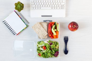 Sebzeler ve beslenme çantasında taze yeşil salata. Çalışma masasında, dizüstü bilgisayar ve diğer ofis malzemeleriyle birlikte. İş yerinde sağlıklı bir öğle yemeği..