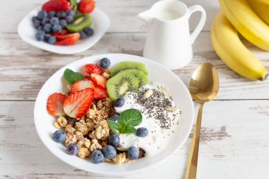 Beyaz seramik kase ev yapımı granola yoğurt, taze böğürtlen ve beyaz ahşap masa arkasında meyveler. Sağlıklı yeme kavramı.